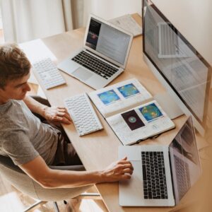 A man multitasks with laptops and a desktop, coding in a home office setting.